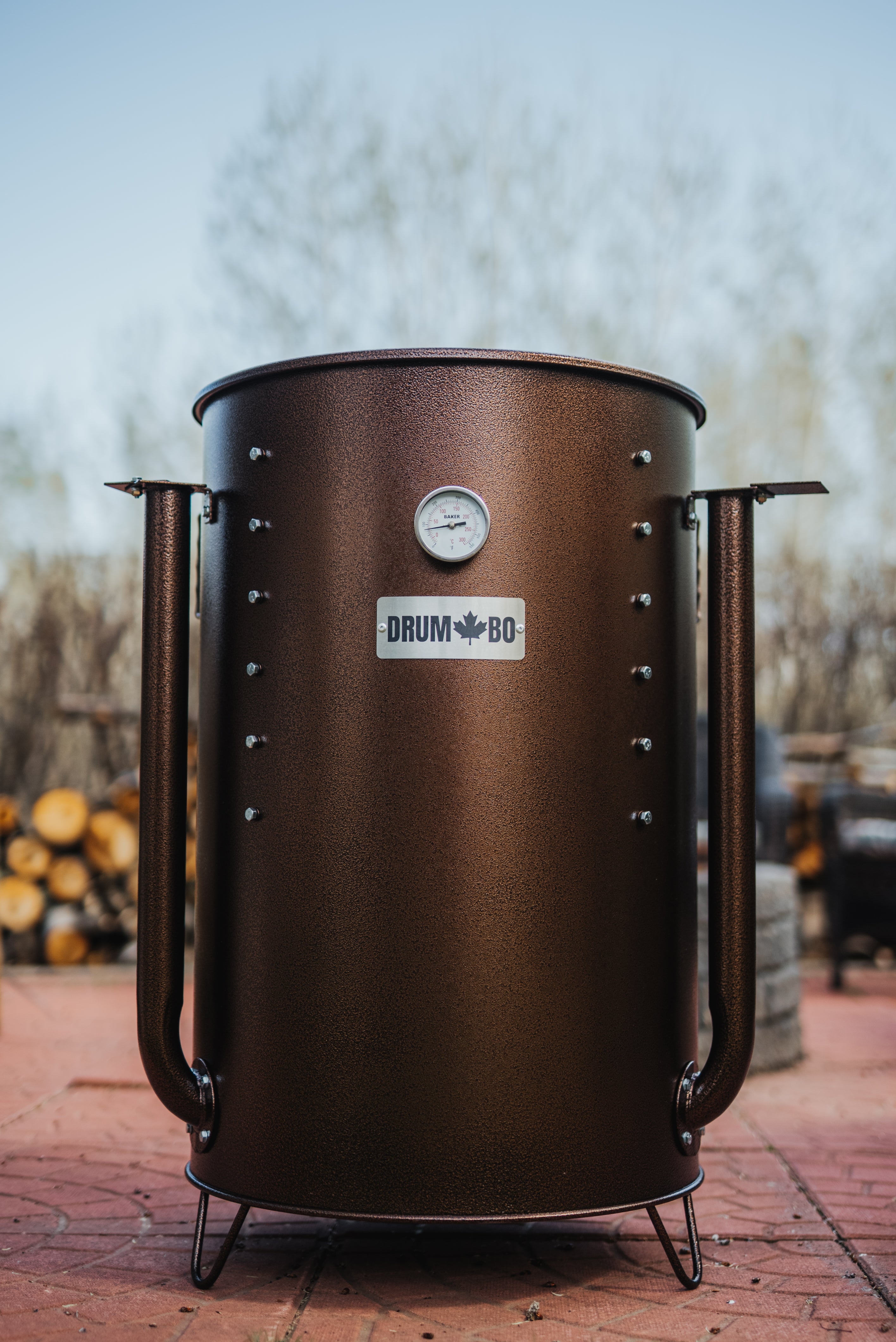 copper metal drum smoker with 'DRUMBO' branding on a brick surface.